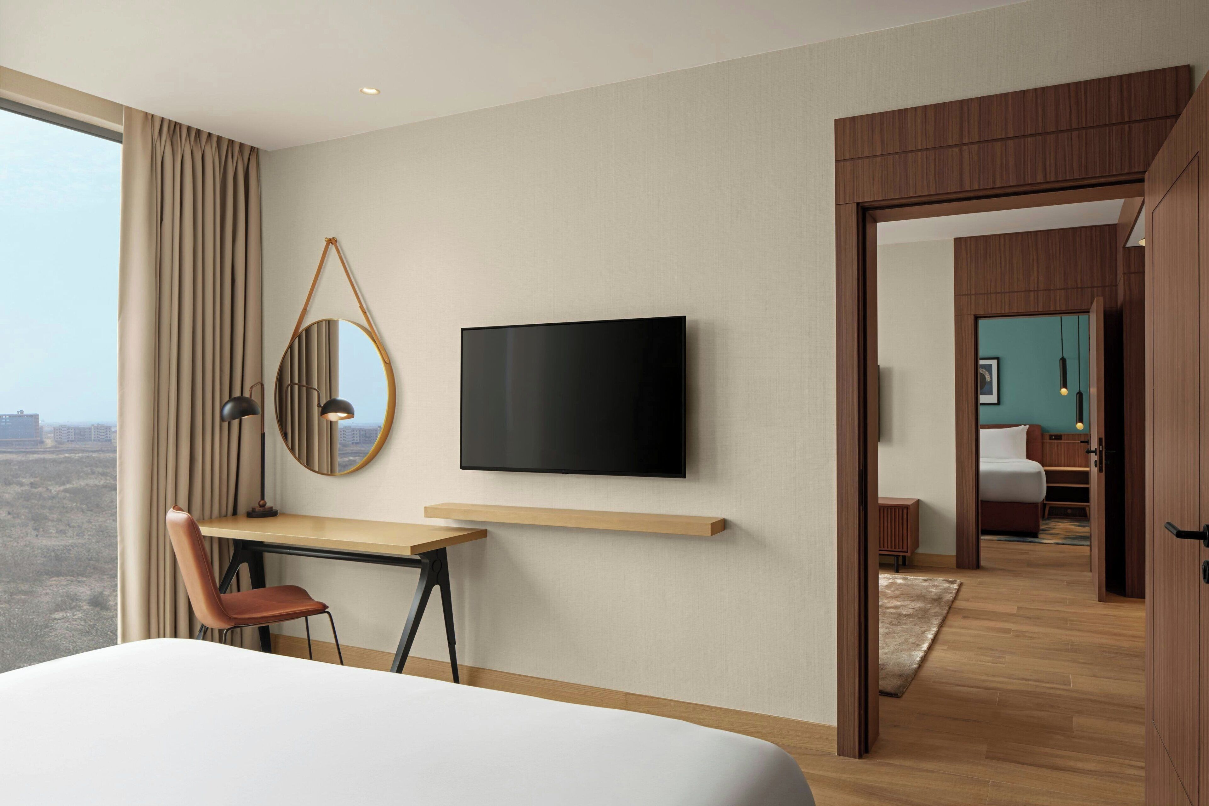 A hotel room with a bed, a desk with a chair, a TV mounted on the wall, and a view of the city through large windows.