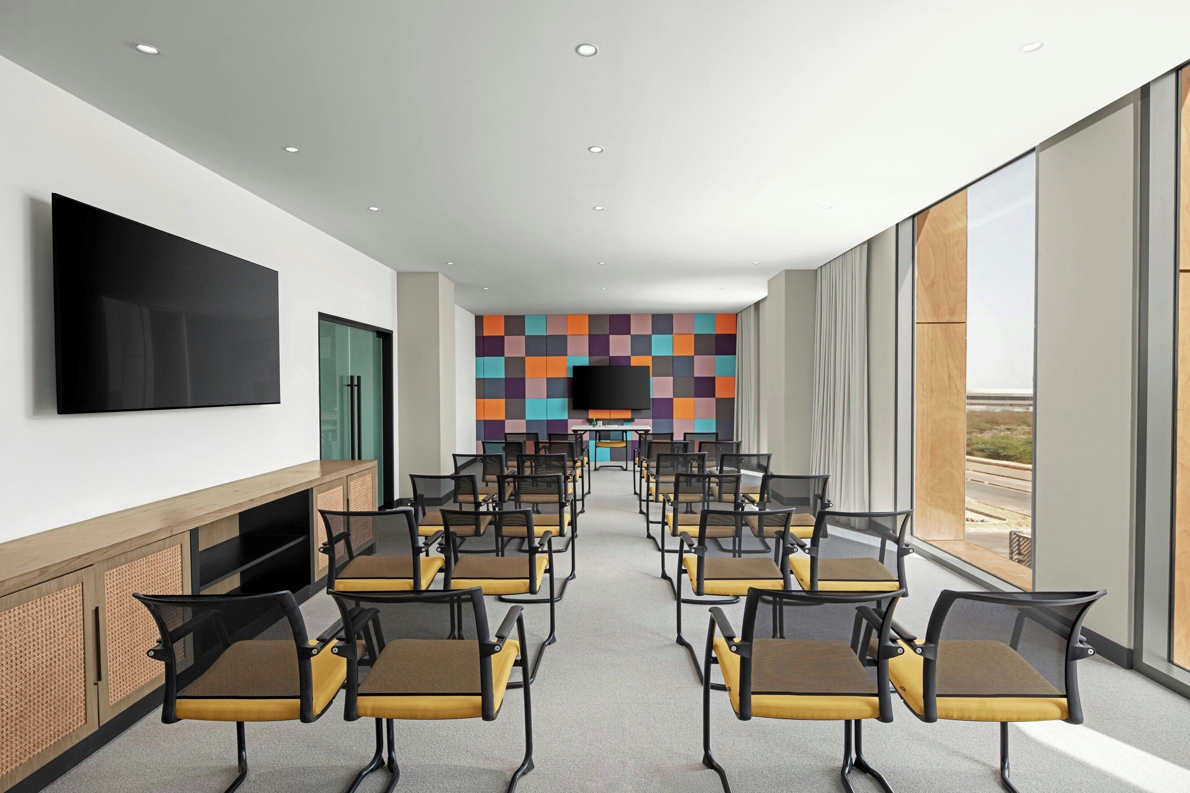 A conference room with rows of chairs, a large flat-screen TV on the wall, and a colorful wall design.