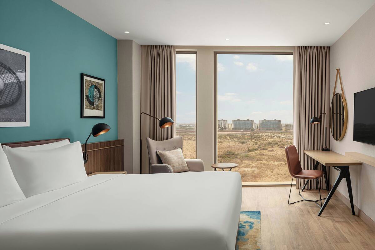 A hotel room with a large bed, a desk, a chair, and a TV. There is a window with a view of buildings and a landscape.