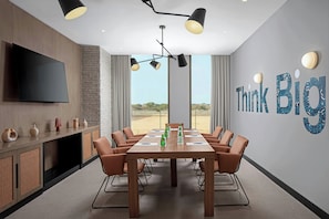 Meetings take place around a long table surrounded by six comfortable chairs, with a large screen and a window offering a view of the outdoors.
