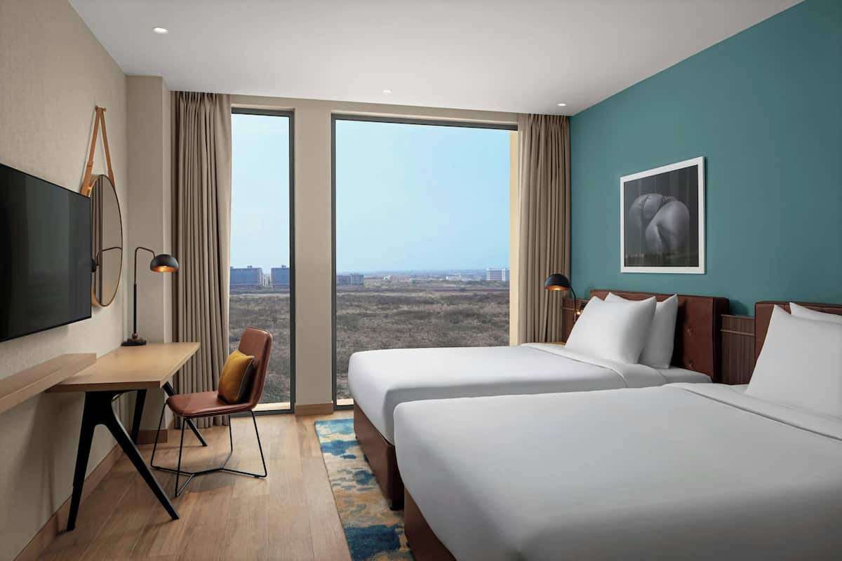 A hotel room with two beds, a desk, a chair, a TV, and a large window with a city view.