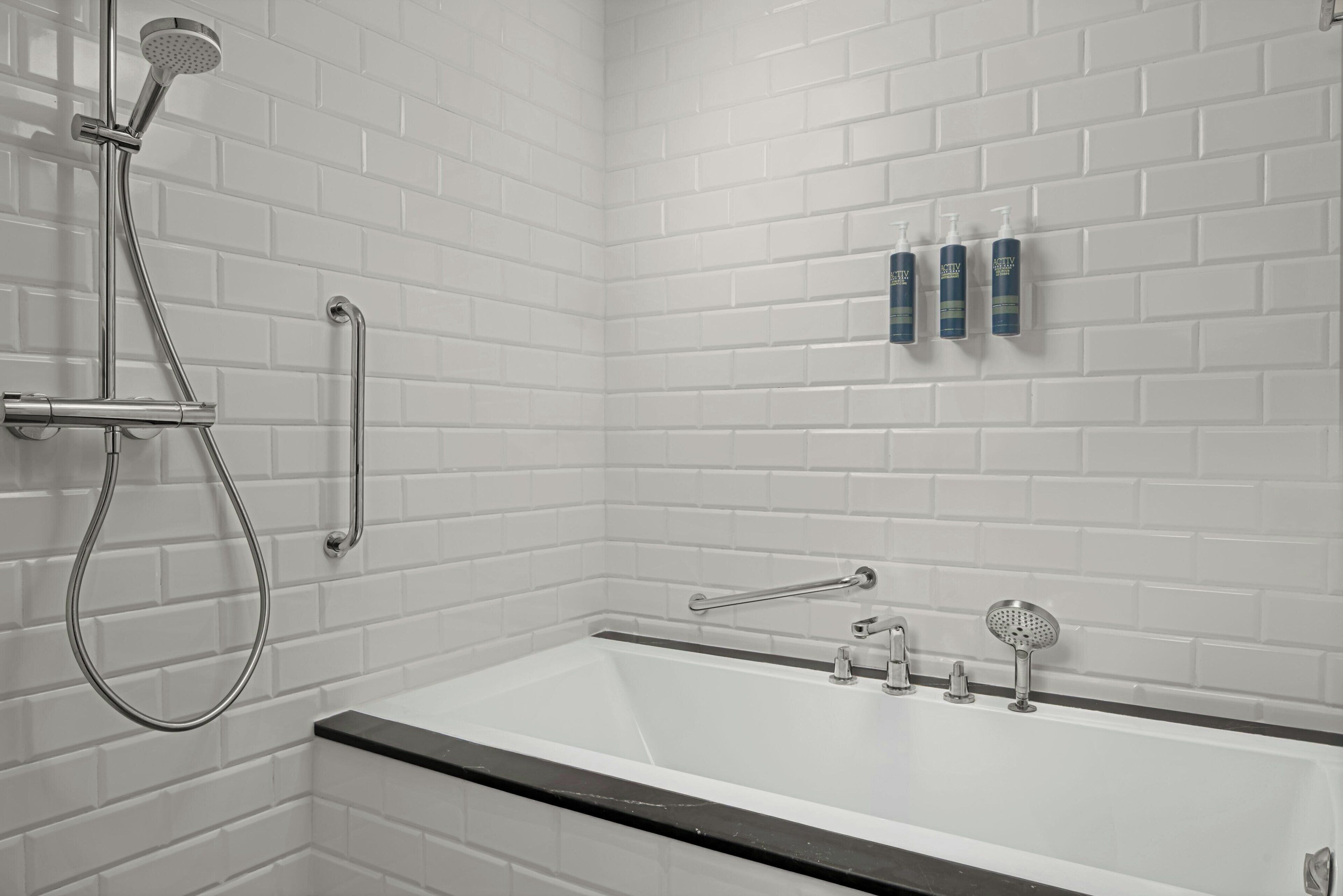 Un bagno moderno con parete rivestita in piastrelle bianche, una vasca da bagno, un soffione a mano e una barra di sostegno.