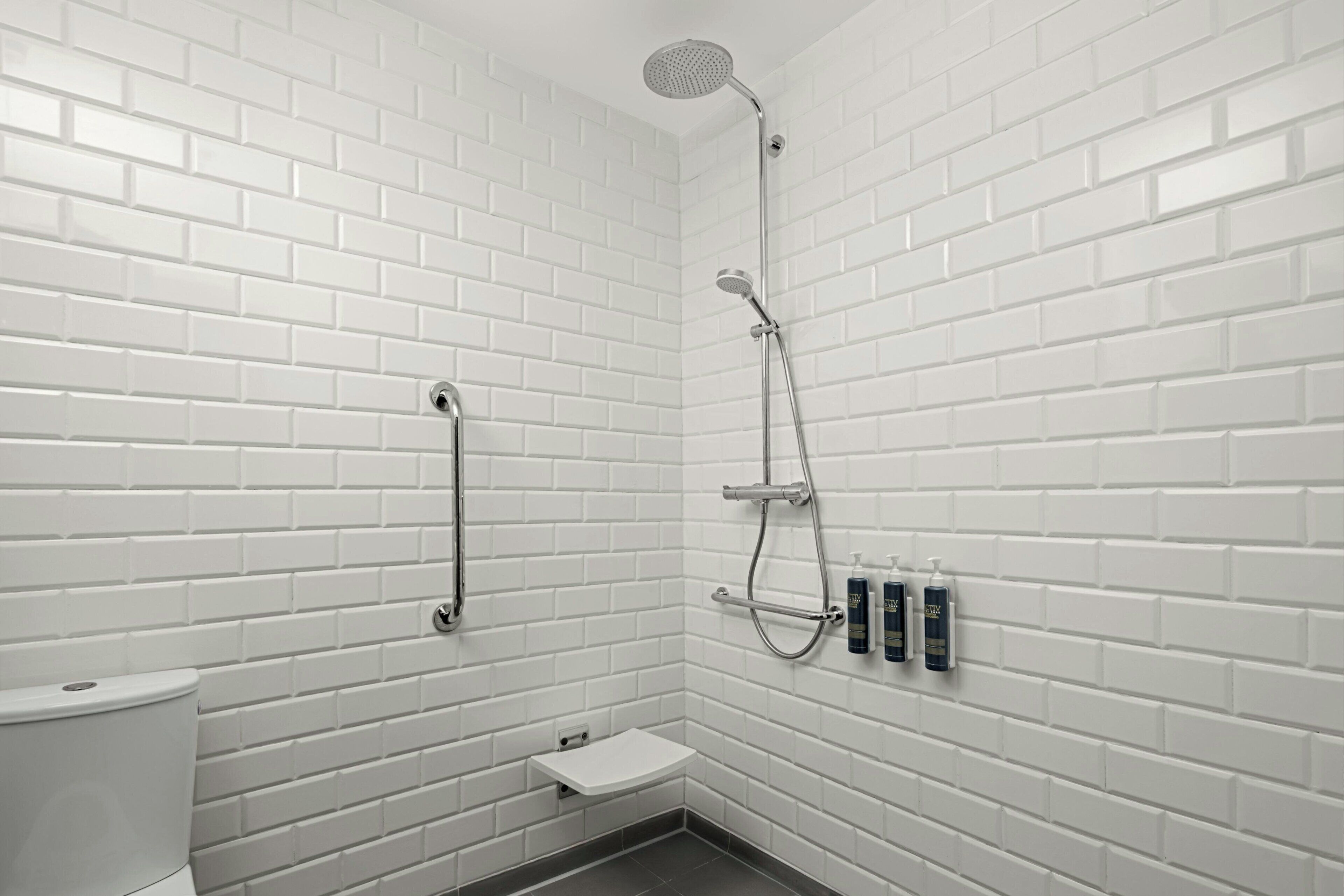 Une petite salle de douche aux murs recouverts de carreaux blancs, équipée d’un pommeau de douche à main et de deux bouteilles de shampoing fixées au mur.