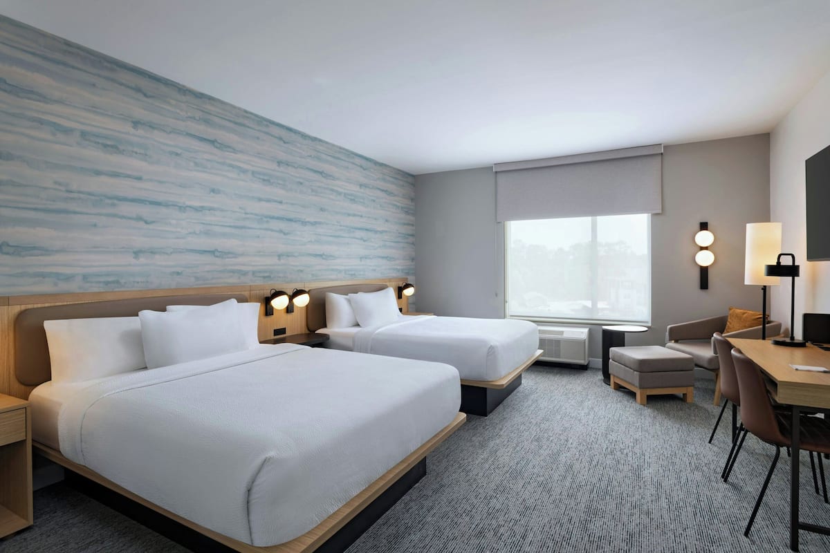 A hotel room with two beds, a desk, a chair, a lamp, and a large window.