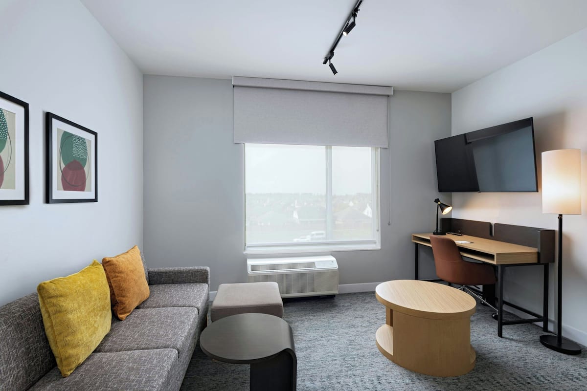 A modern hotel room with a grey sofa, a small round table, a desk with a chair, a flat-screen TV, and a large window with a view.