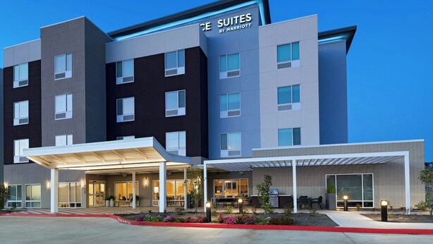 A modern hotel with a sign reading "The Suites by Marriott." It features a multi-story building with a covered entrance and outdoor seating area. - TownePlace Suites by Marriott® Houston Tomball (Tomball)