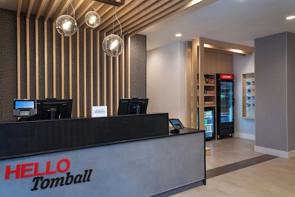 A hotel reception desk with a 'HELLO Tomball' sign, a self-service kiosk, a refrigerator stocked with beverages, and a 'Just chillin'' sign. - TownePlace Suites by Marriott® Houston Tomball (Tomball)