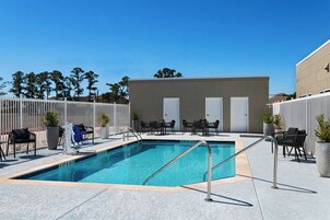 Una piscina con una escalera metálica, rodeada de sillones y mesas, con un edificio al fondo.