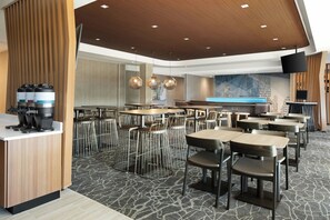 A modern dining area with multiple tables and chairs, a kitchen counter with coffee machines, and a large screen displaying text.