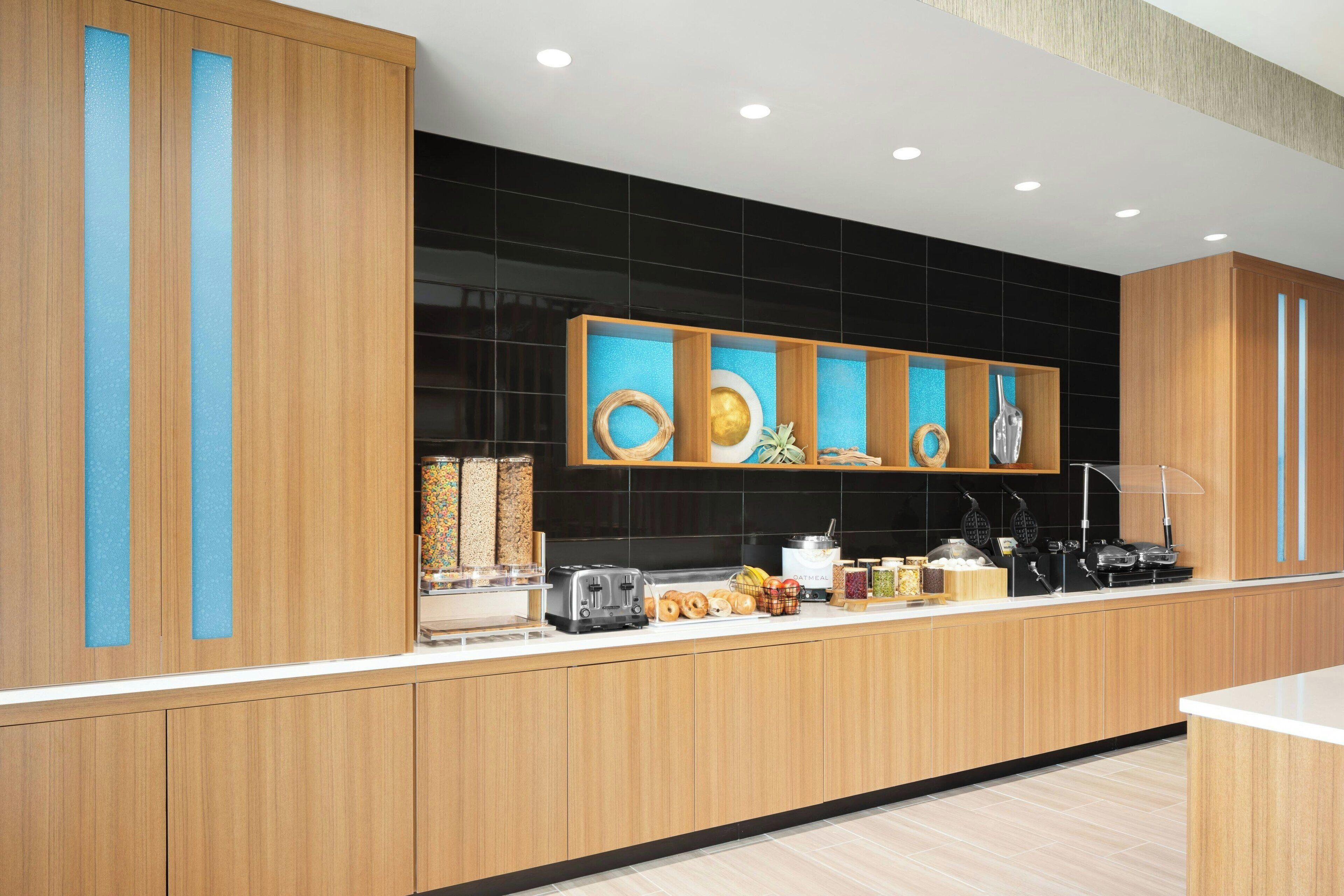 A modern kitchen with wooden cabinets, a black backsplash, and a countertop with various kitchen appliances and food items.