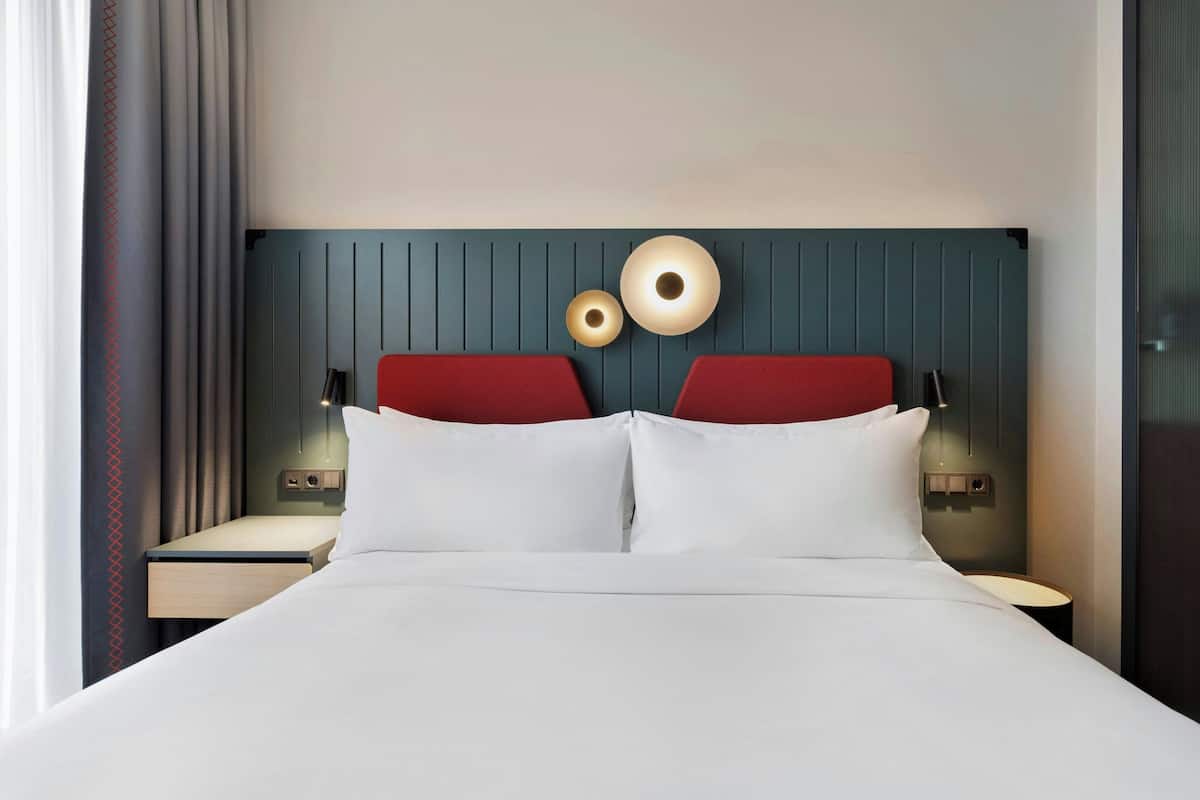 A hotel room with a bed, two red headboards, bedside lamps, and a wall-mounted light fixture.