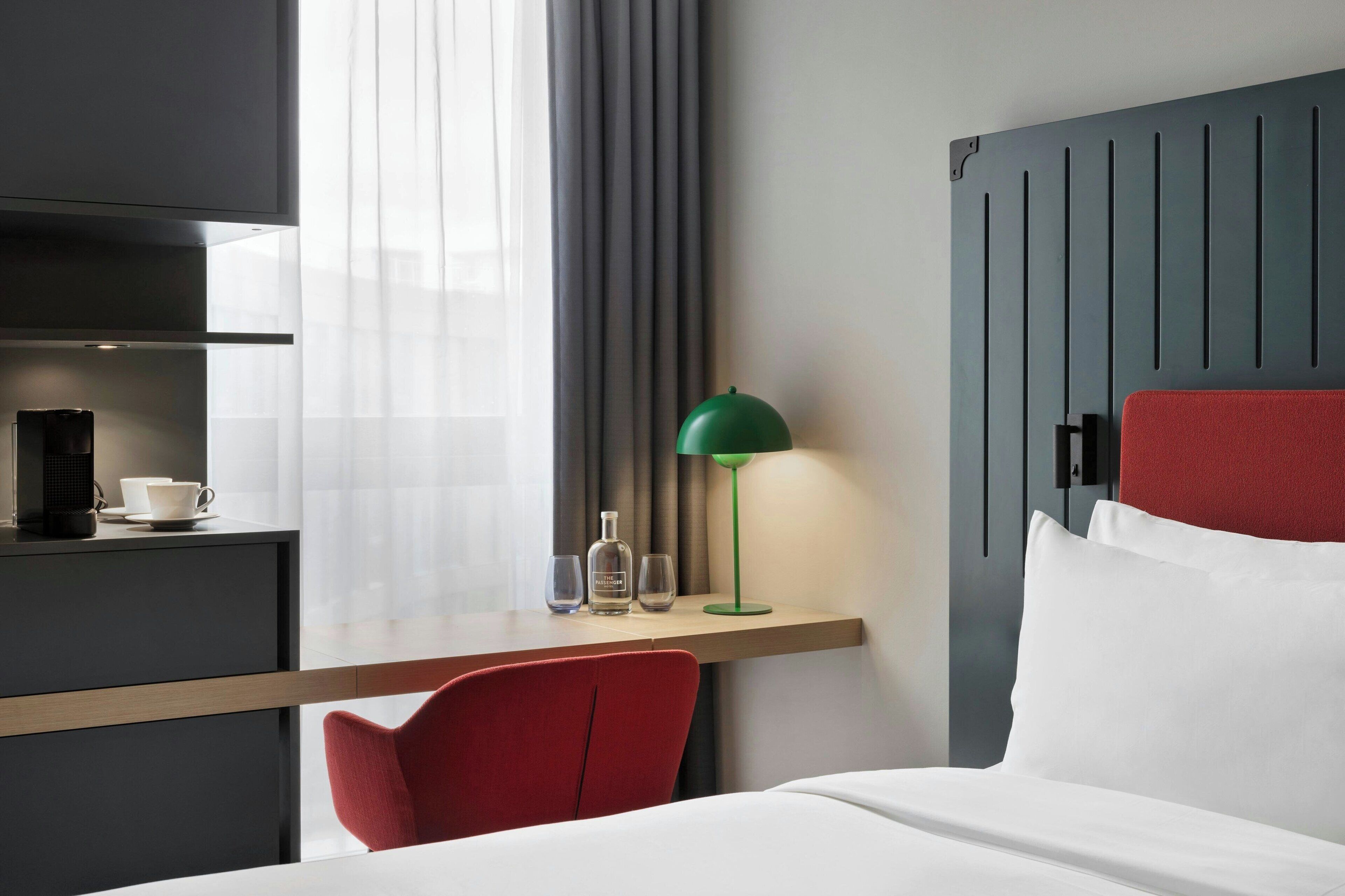 Una habitación de hotel con una cama, una silla roja, un escritorio con lámpara, una cafetera y una botella.