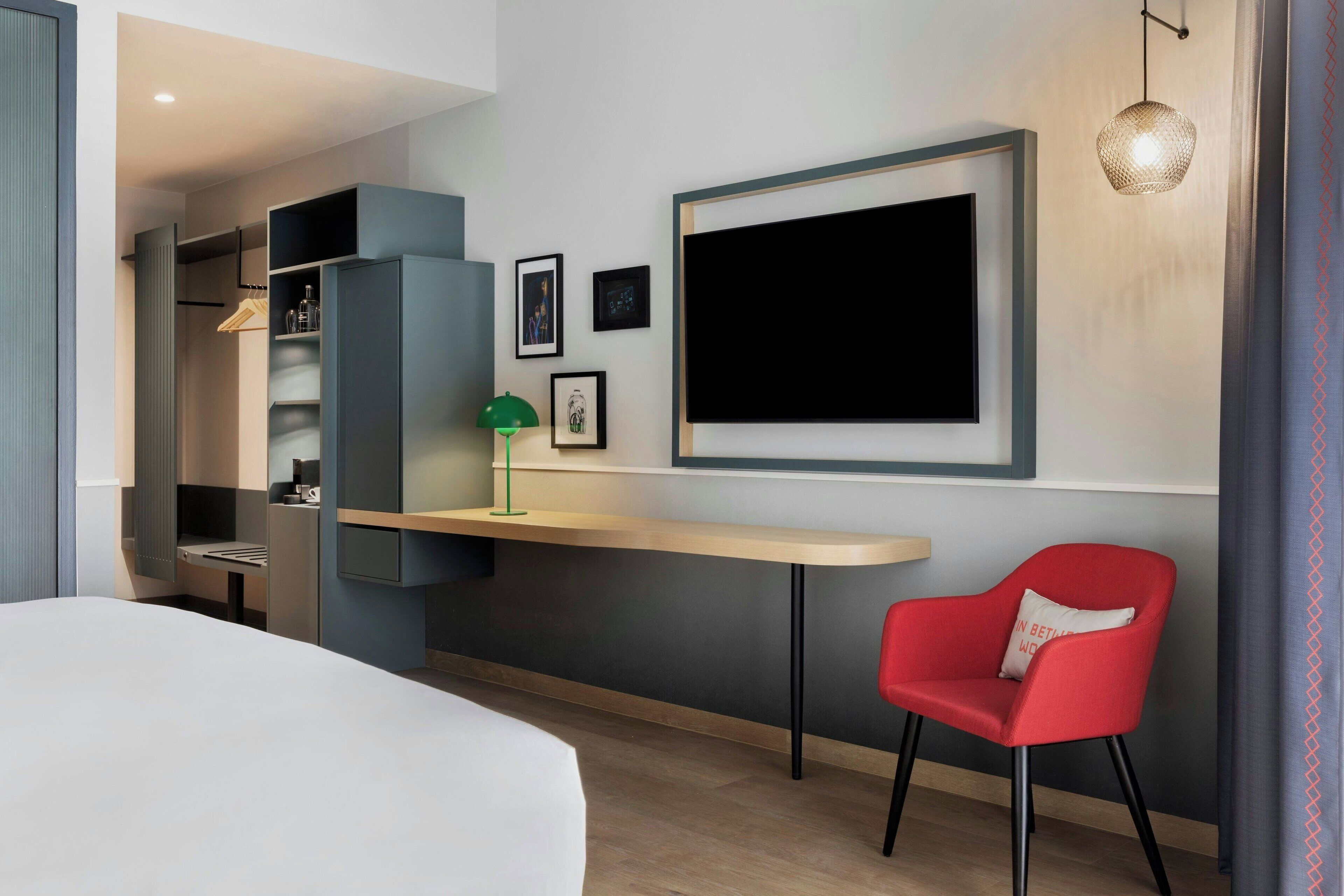 A modern hotel room with a bed, a desk, a red chair, and a large TV on the wall.