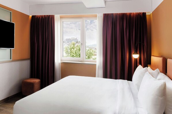 A hotel room with a bed, a window with curtains, a flat-screen TV, and a lamp. - The Passenger, a Tribute Portfolio Hotel (Salzburg)