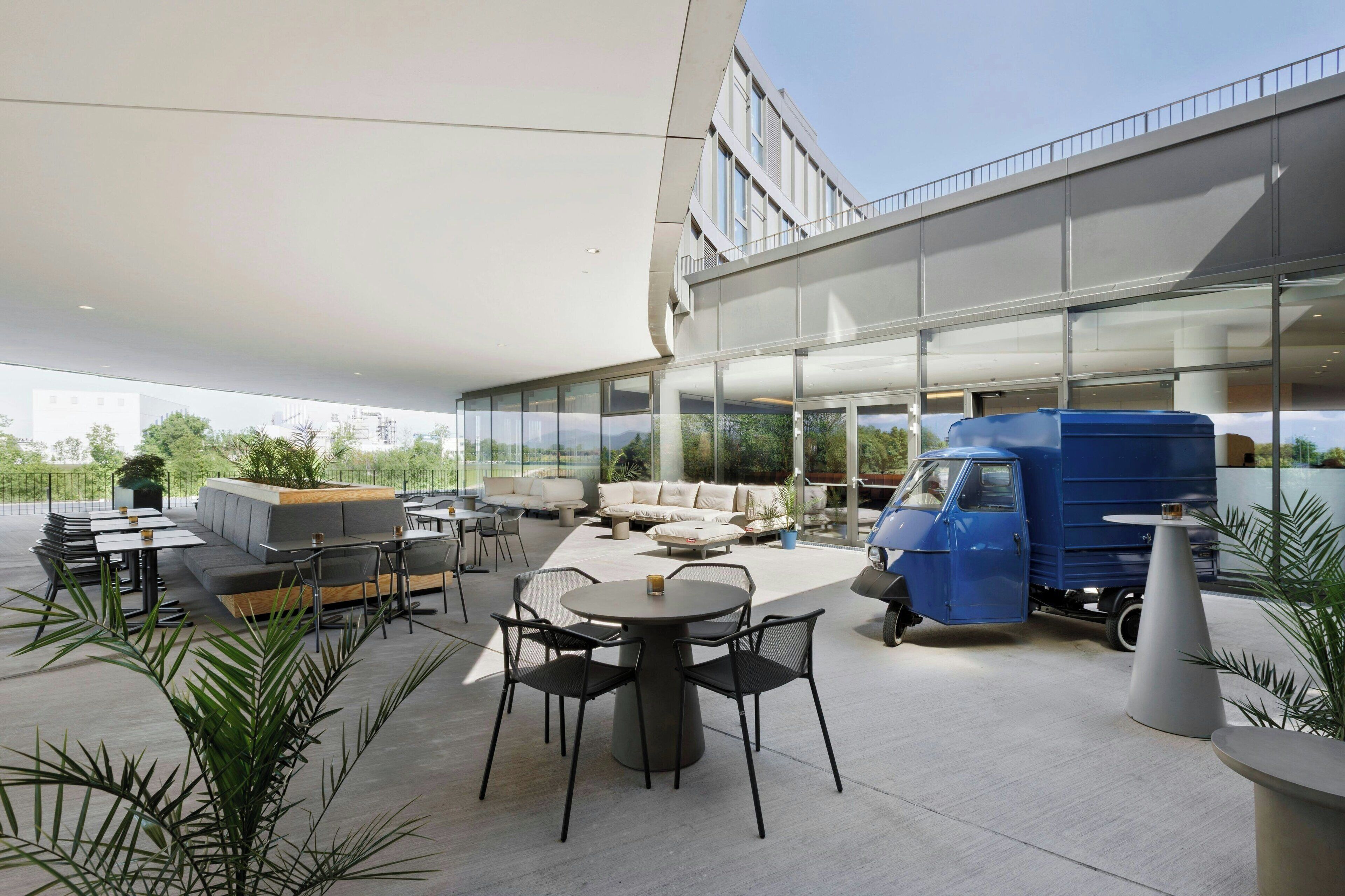 An outdoor seating area with a round table and chairs, a blue delivery truck, and a modern building with large windows.