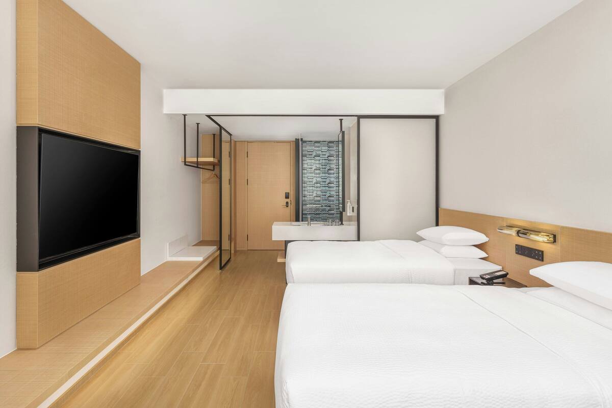 A modern hotel room with a large flat-screen TV, two beds with white bedding, a wooden floor, and a wall-mounted light fixture.