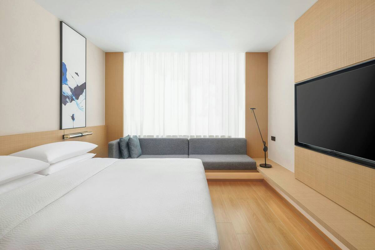 A modern hotel room with a large bed, a sofa, a TV, and a window with sheer curtains.