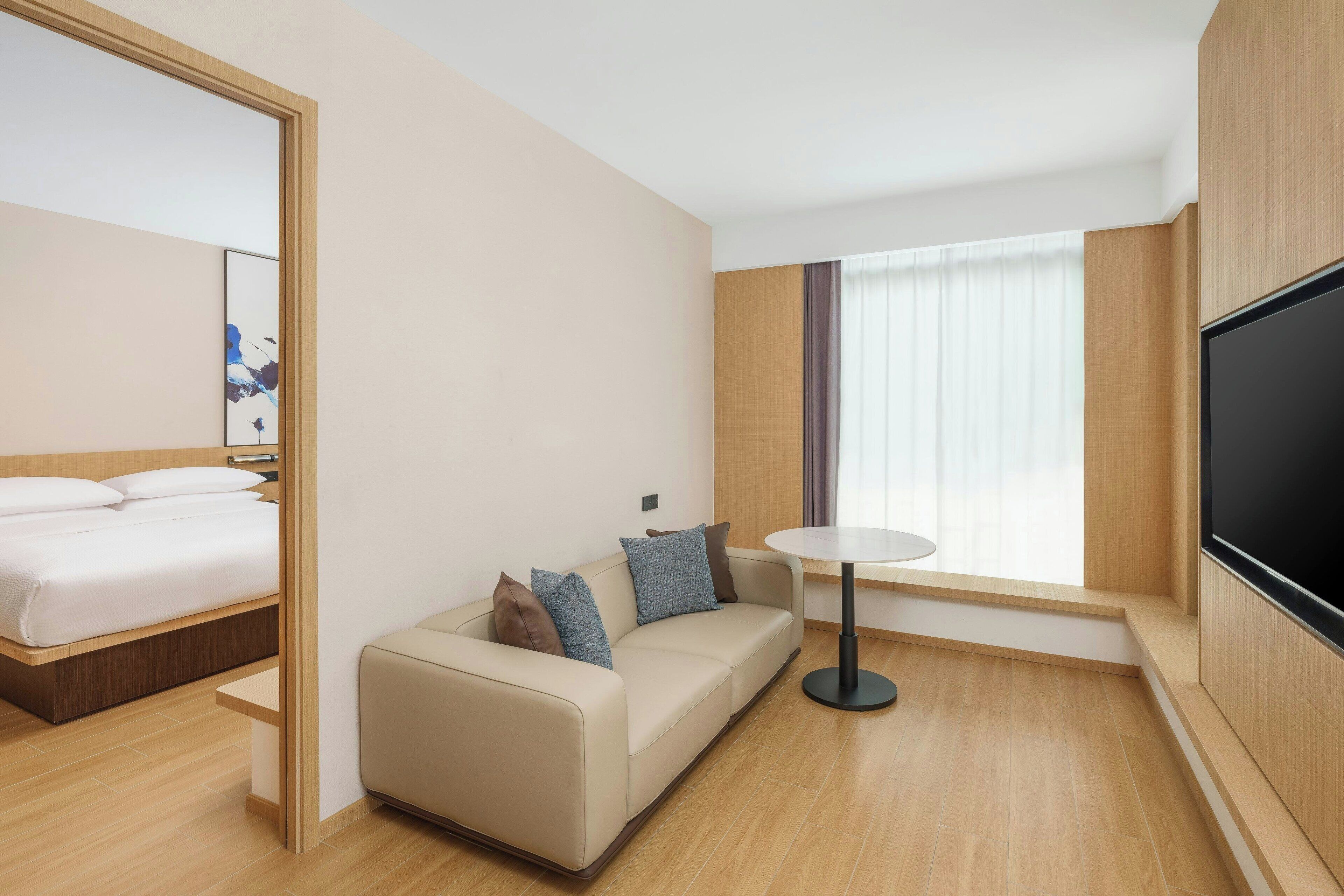 A modern hotel room with a bed, a sofa, a TV, and a window with curtains.