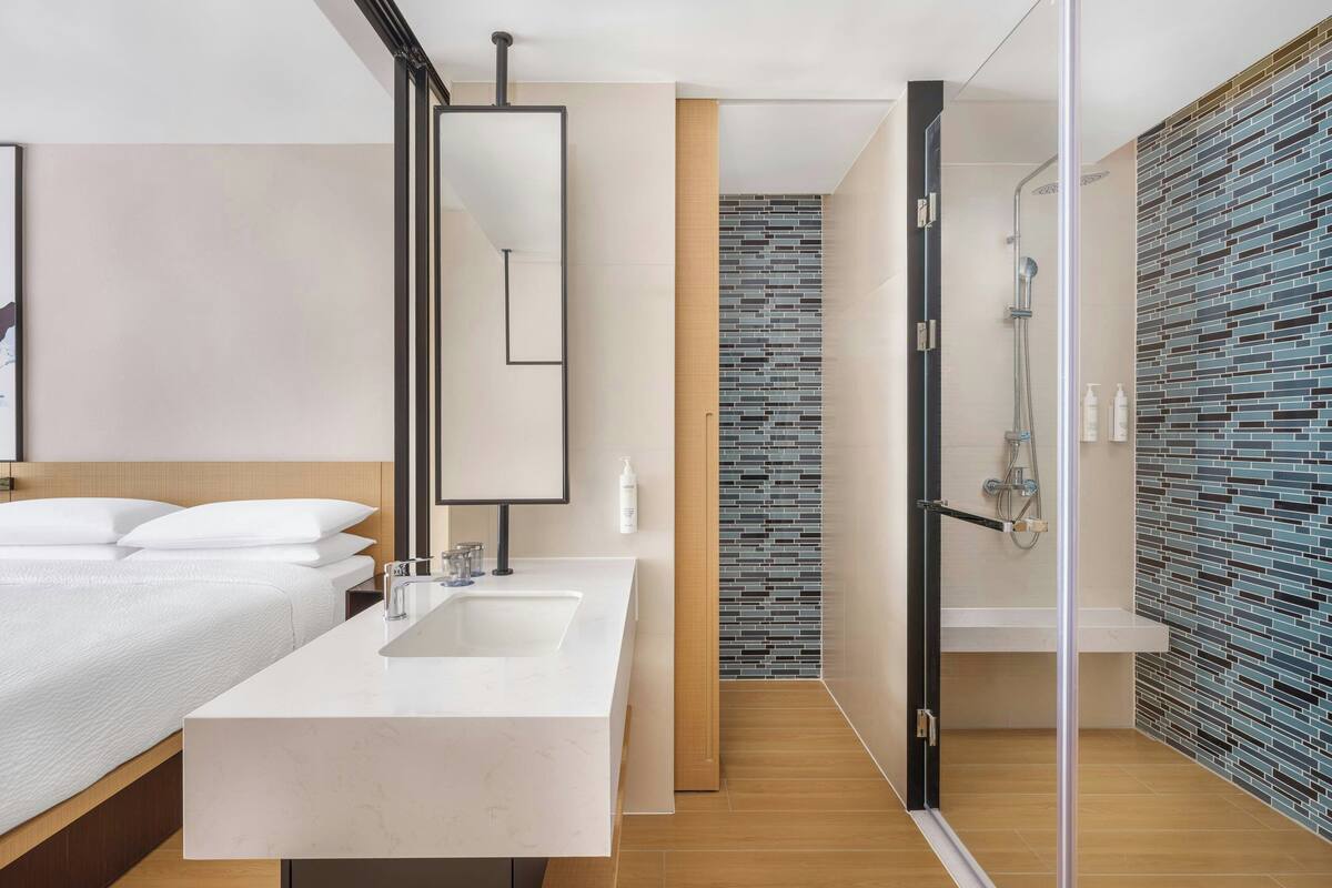 A modern hotel room with a large bed, a bathroom with a glass shower enclosure, and a tiled wall.