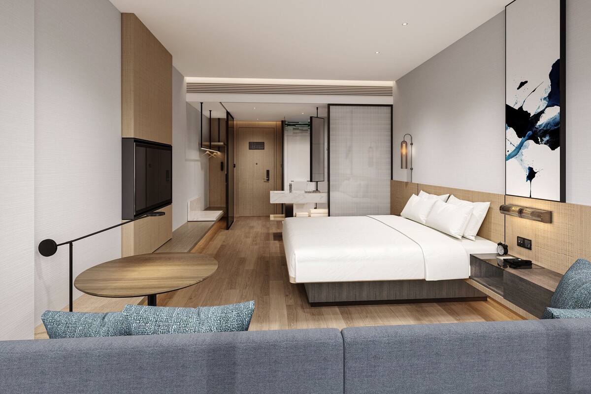 A modern hotel room with a large bed, a sofa, a TV, and a bathroom visible through a glass door.