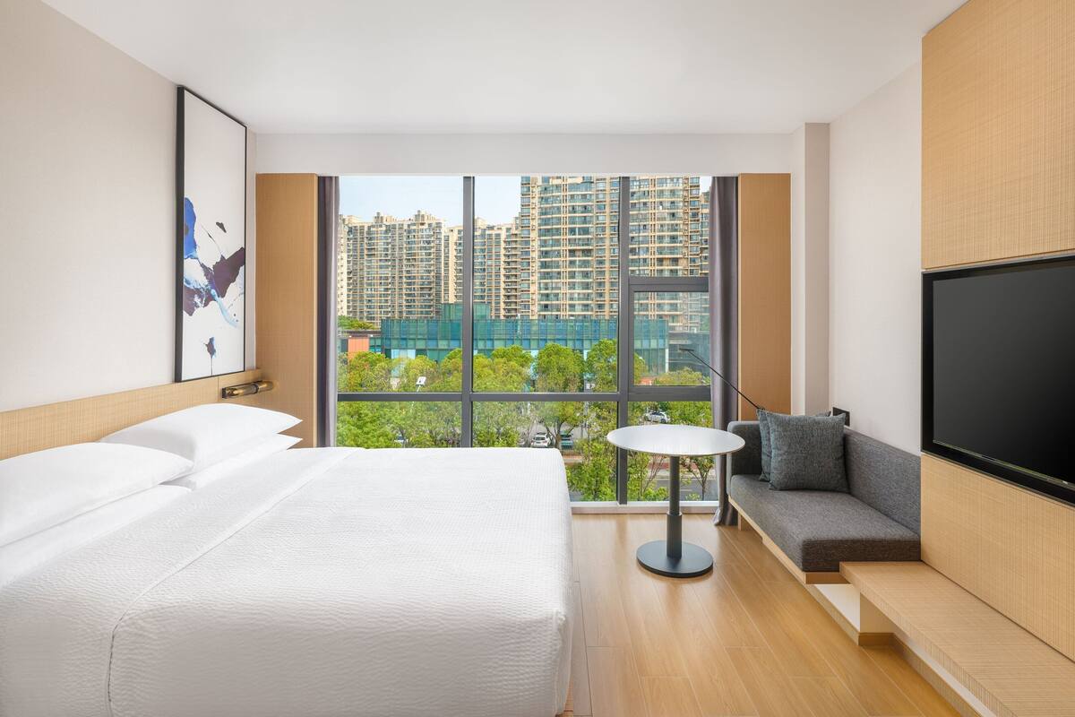A hotel room with a bed, a sofa, a TV, and a large window offering a view of high-rise buildings and greenery.