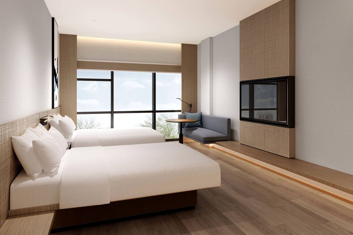 A modern hotel room with a large bed, a sofa, a TV, and a large window offering a view of the sky.