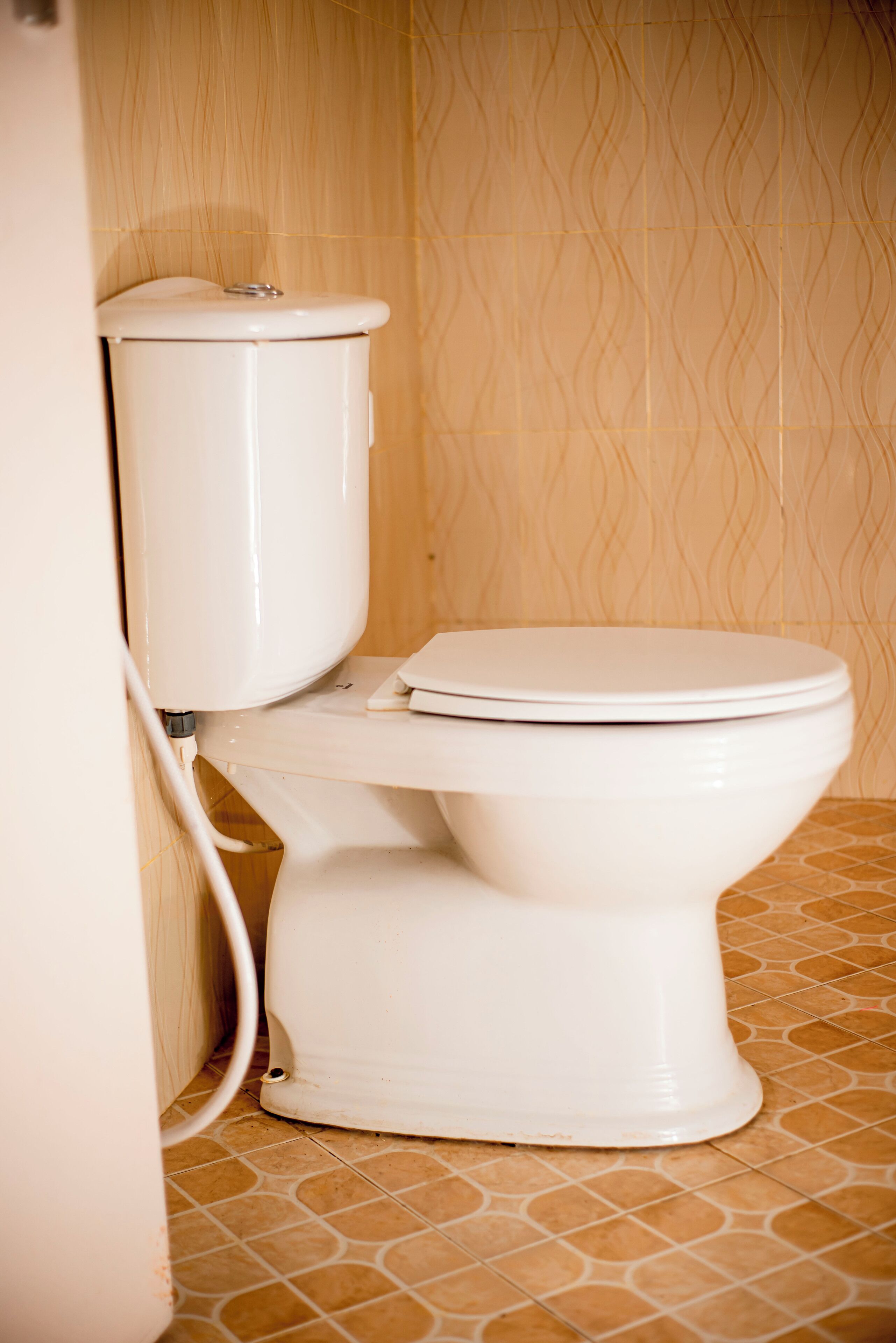 Et hvidt toilet med tank og låg, placeret i et hjørne med fliserede vægge og gulv.