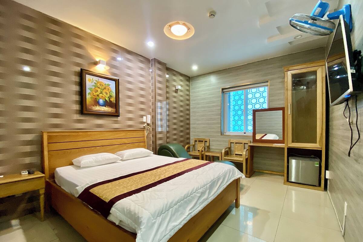 Ein Hotelzimmer mit einem hölzernen Bett, einem Schreibtisch, einem Stuhl, einem kleinen Tisch, einem an der Wand befestigten Fernseher und einem Fenster mit geometrischem Muster.