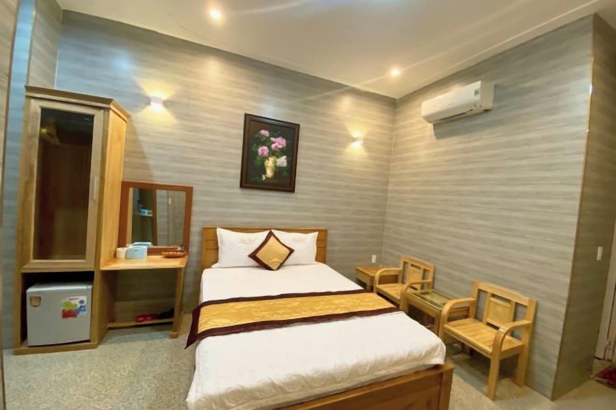 A bedroom with a bed, a wooden wardrobe, a small table, two chairs, and an air conditioning unit on the wall.