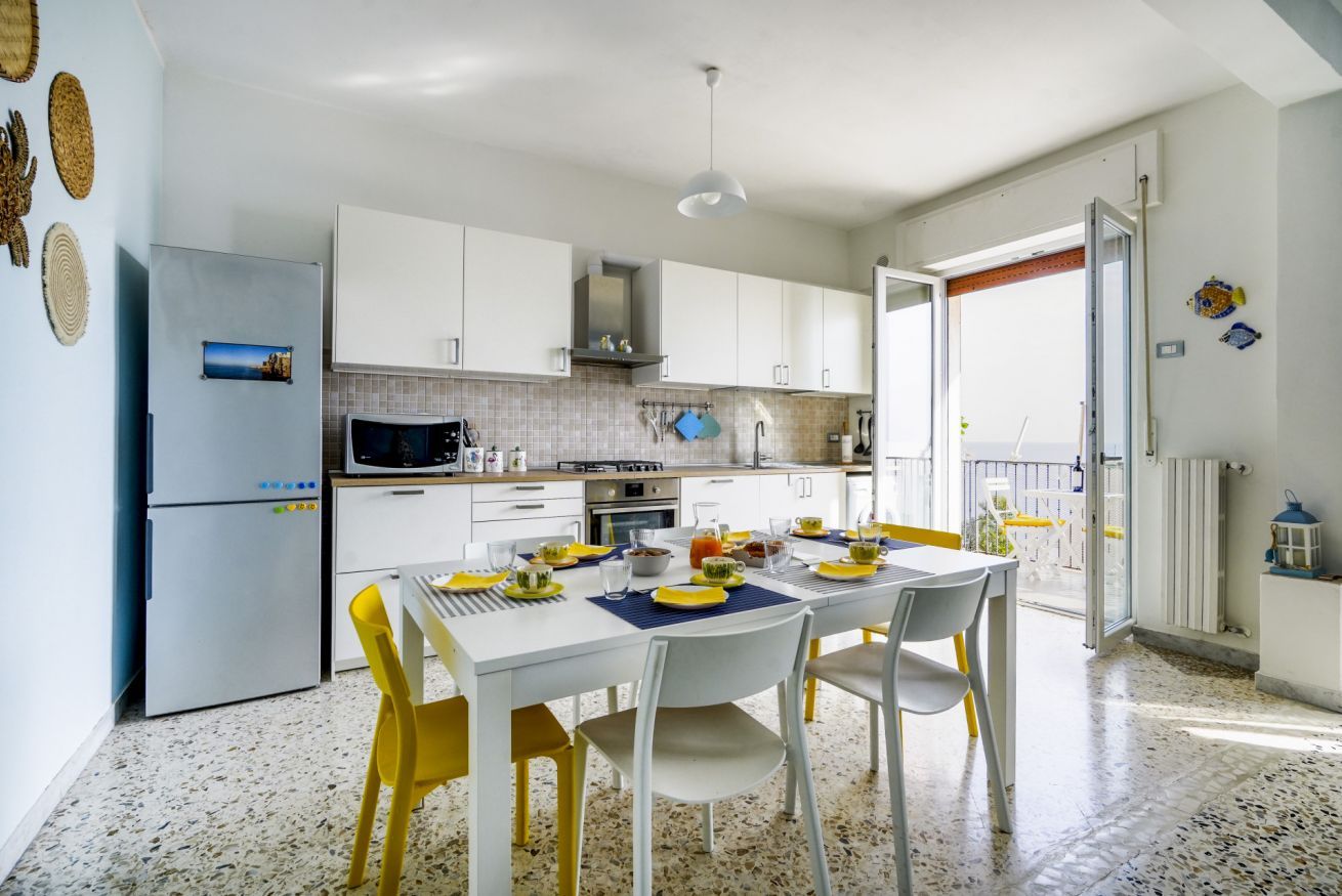 A modern kitchen with white cabinets, a dining table set with yellow chairs, and a view through an open door to an outdoor area.