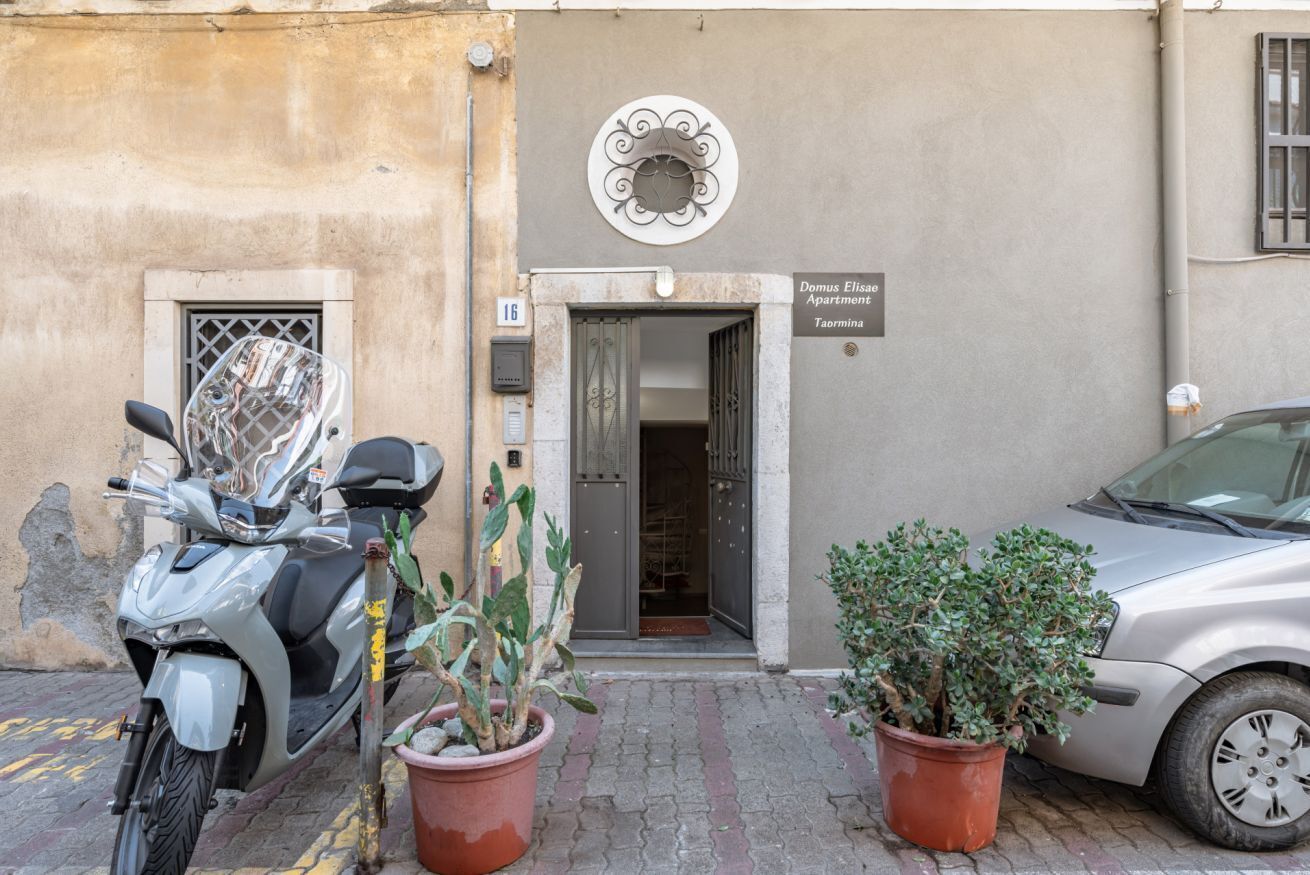 Entrada de un edificio con un cartel que dice 'Domus Elisio Apartment Taormina', una scooter estacionada y dos macetas.