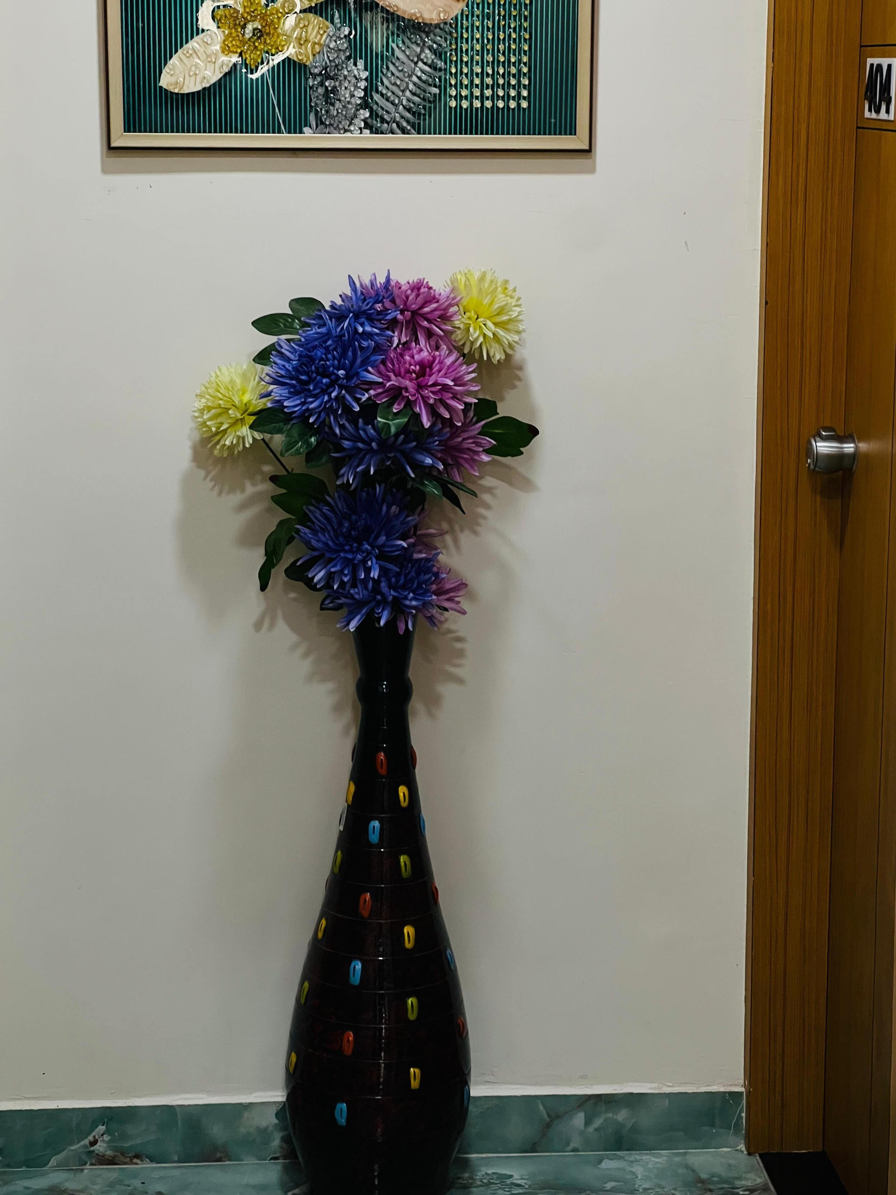 Eine Vase mit bunten Blumen auf einem Fliesenboden, eine Tür und ein gerahmtes Bild an der Wand.