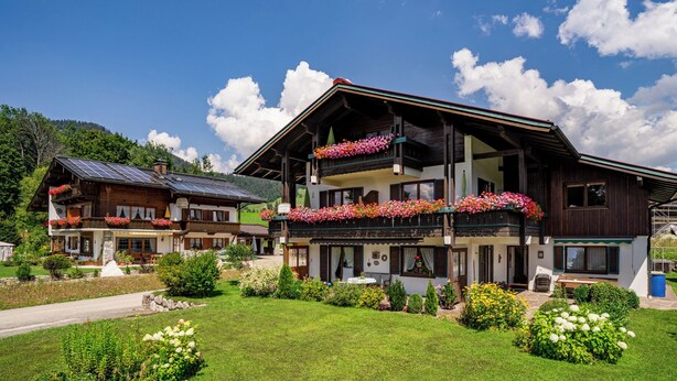 Un bâtiment en bois à deux étages, doté de balcons ornés de fleurs, entouré de jardins bien entretenus et surplombé d’un ciel dégagé.