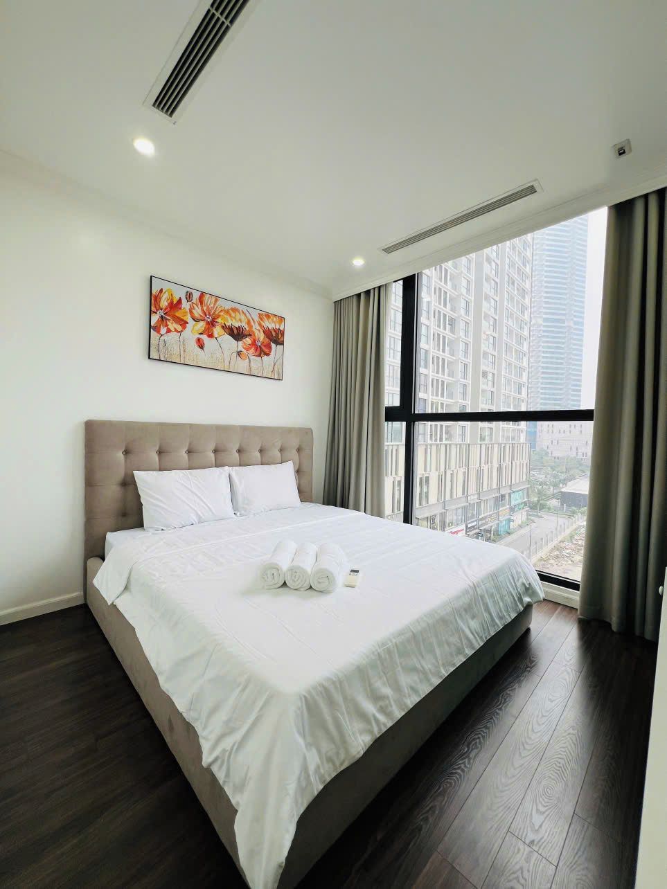 A modern hotel room with a large bed, a city view, and a floral painting on the wall.