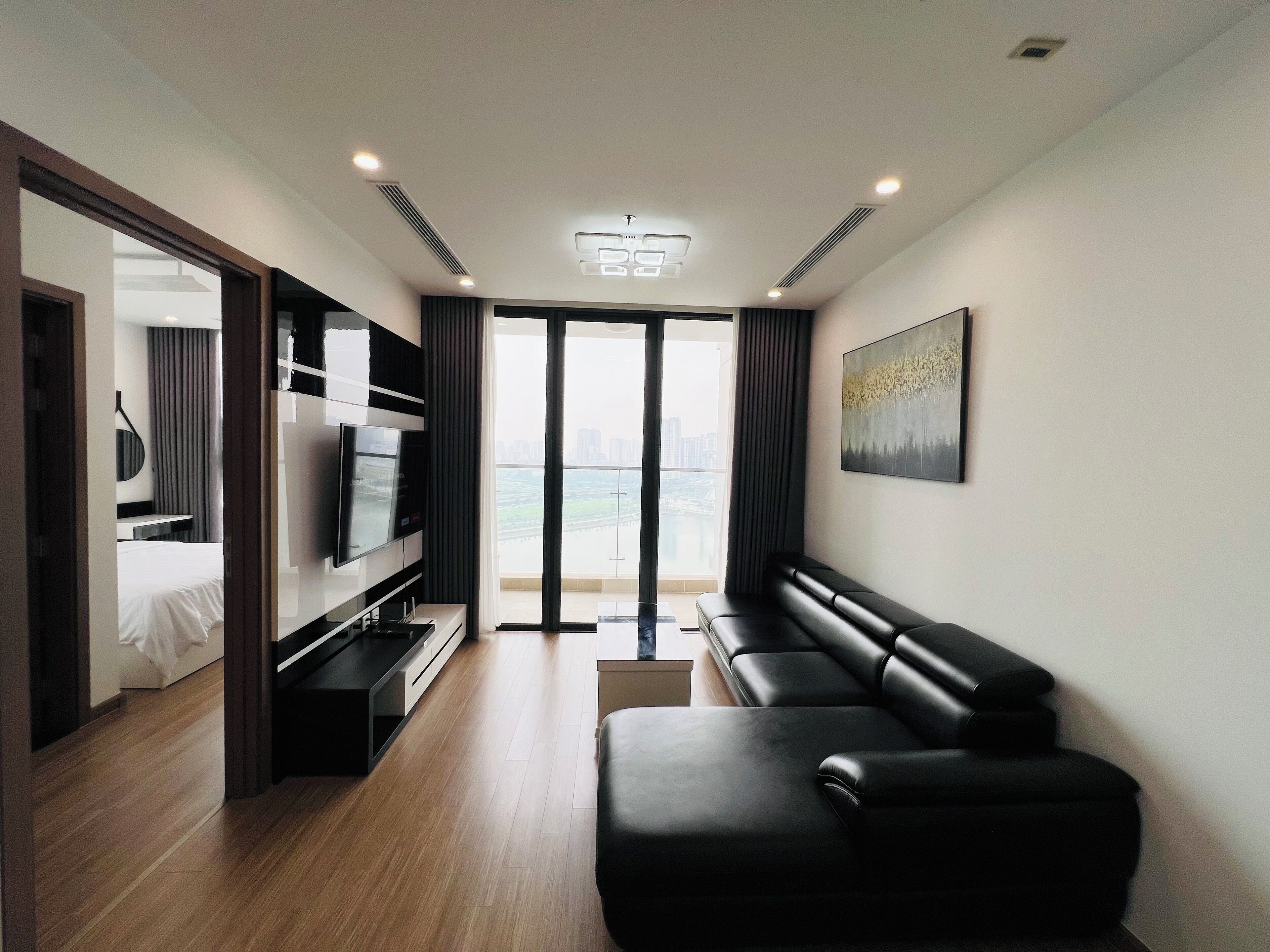 A modern hotel room with a large flat-screen TV, a black leather sofa, a dining table, and a view of the city through large windows.