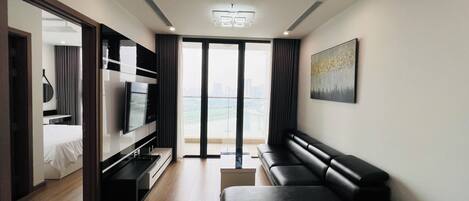A modern hotel room with a large flat-screen TV, a black leather sofa, a dining table, and a view of the city through large windows.