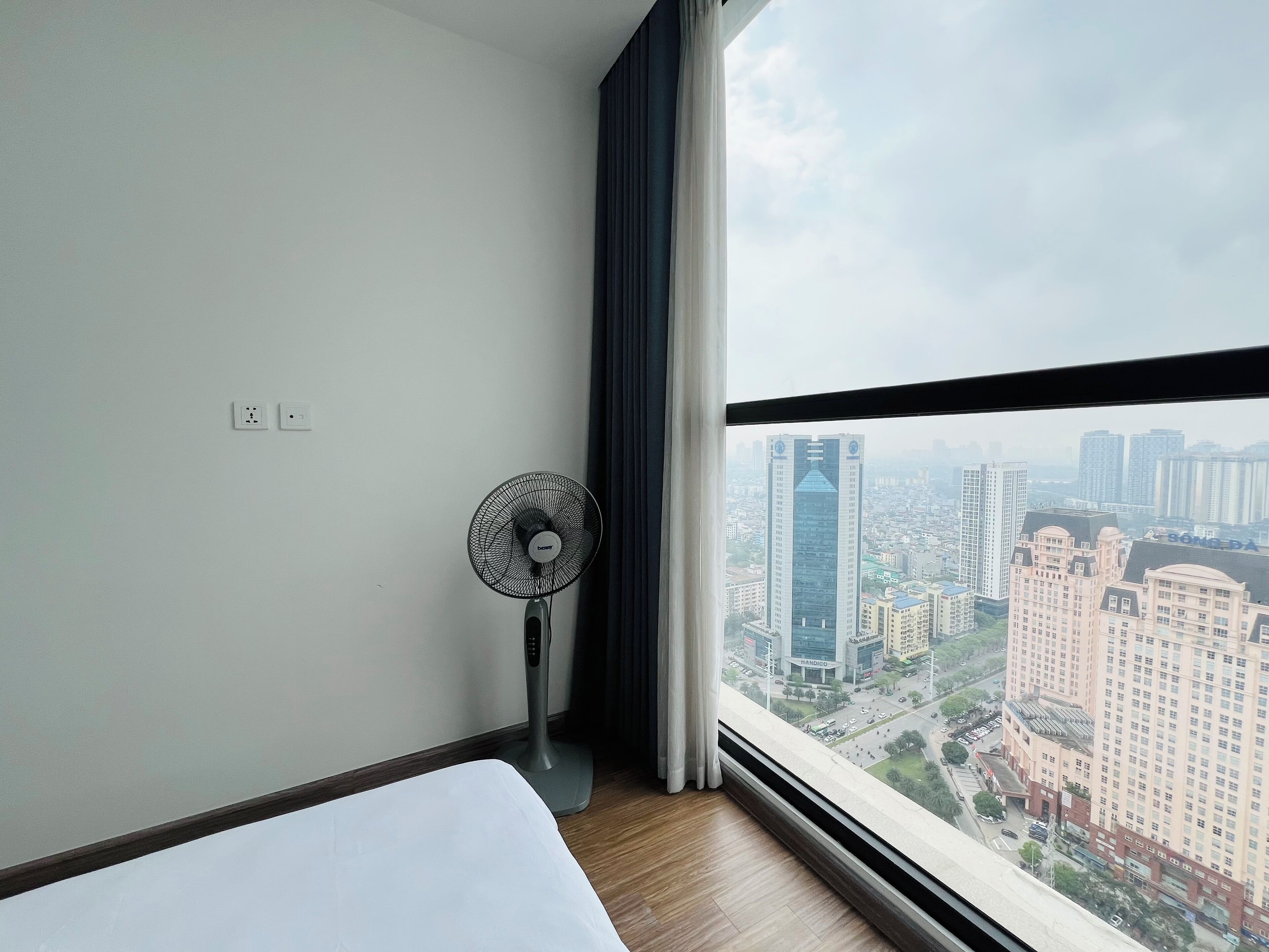 Ein Hotelzimmer mit Bett, einem Standventilator, einem Fenster mit Blick auf die Stadt und einem Vorhang.