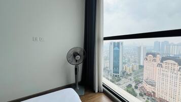 Ein Hotelzimmer mit Bett, einem Standventilator, einem Fenster mit Blick auf die Stadt und einem Vorhang.
