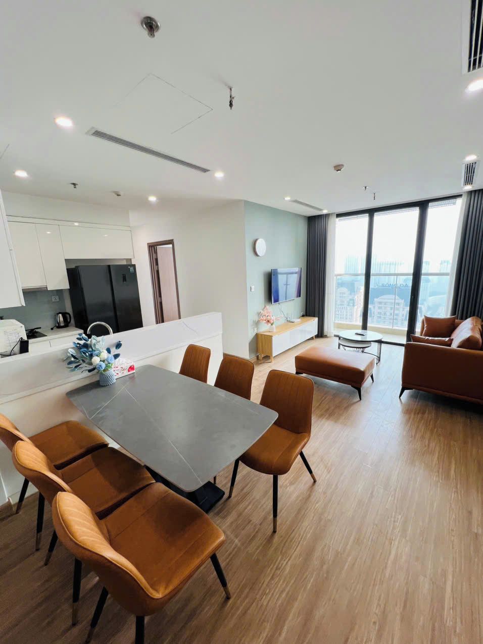 A modern living room with a dining area, a kitchen, and a large window offering a city view.