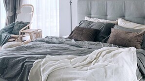 Une chambre à coucher moderne avec un lit recouvert d’un tissu gris, une lampe de chevet et une chaise près de la fenêtre.