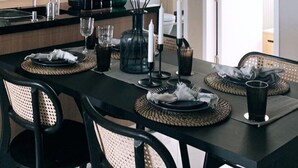 Un espace repas moderne avec une table noire dressée pour quatre personnes, entourée de chaises en osier. Une sculpture originale en papier blanc est suspendue au plafond, et un vase vert contenant des bougies est posé sur la table.