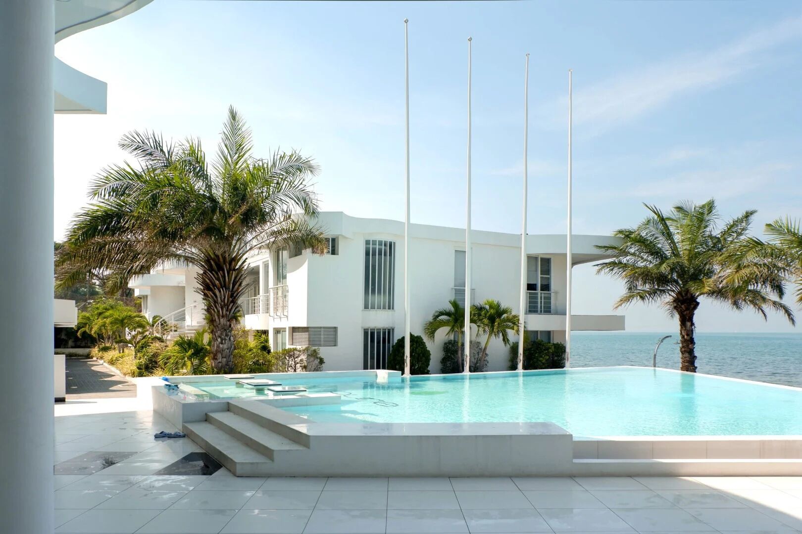 A modern pool area with a clear view of the sea, surrounded by palm trees and a white building.