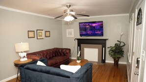 Una sala de estar con un sofá de cuero marrón, un sillón azul, una mesa de centro de madera, una lámpara de pie, un televisor, fotografías enmarcadas en la pared, una chimenea y un ventilador de techo.