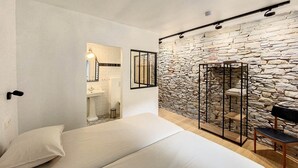 Un dormitorio con una cama blanca, una pared de piedra, un banco de madera y una mesita.
