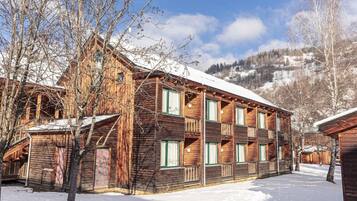 A wooden multi-story building with balconies, surrounded by snow-covered ground and bare trees, set against a mountainous backdrop.