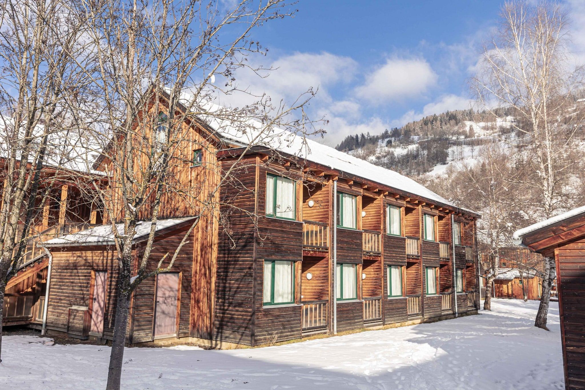 Un edificio de madera de varios pisos con balcones, rodeado de suelo cubierto de nieve y árboles sin hojas, con montañas al fondo.