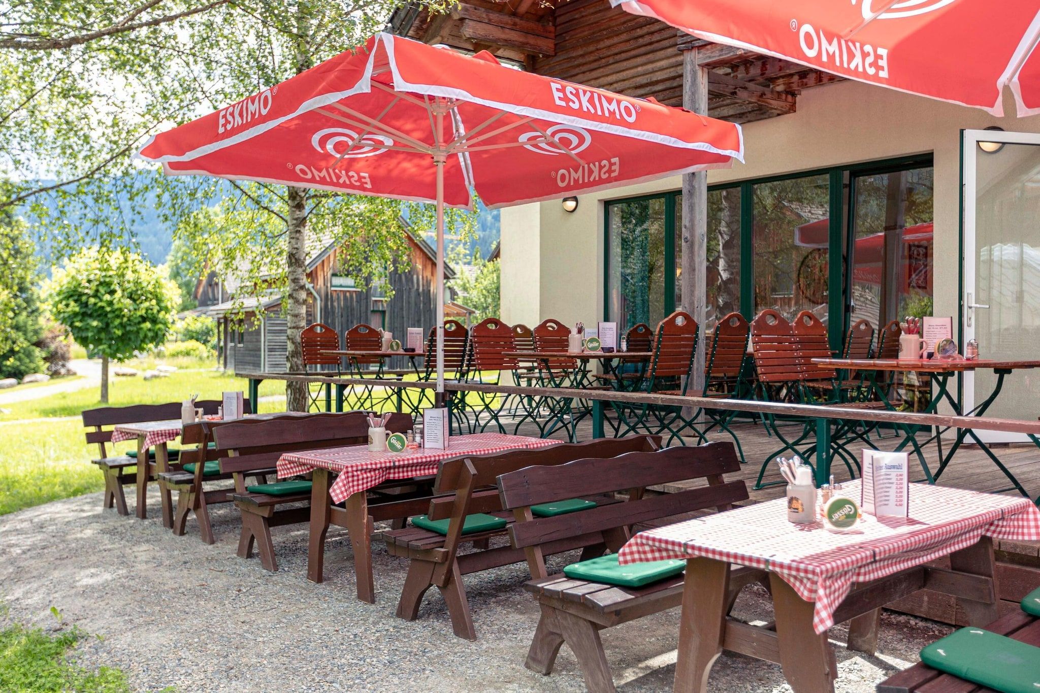 An outdoor dining area with wooden tables and benches, red and white checkered tablecloths, and red umbrellas with the brand name "ESKIMO".