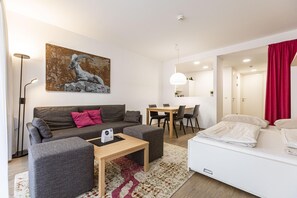 A modern living room with a grey sofa, a small table, a dining area with a white table and chairs, and a bed with white bedding.