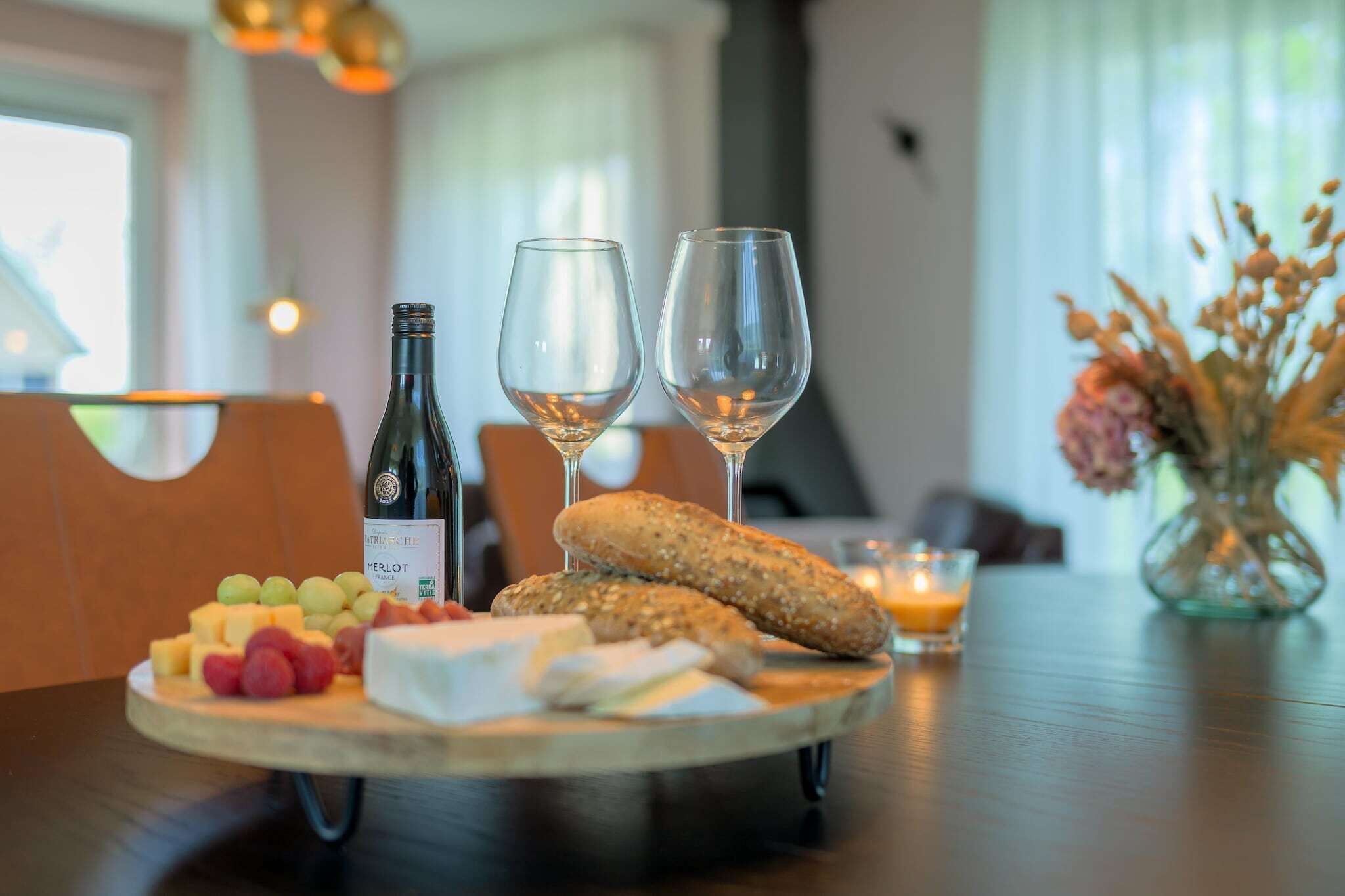 A wooden board with cheese, grapes, a baguette, and two wine glasses on a table.