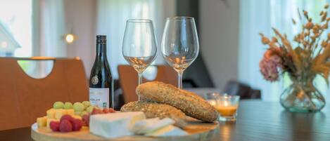 A wooden board with cheese, grapes, a baguette, and two wine glasses on a table.